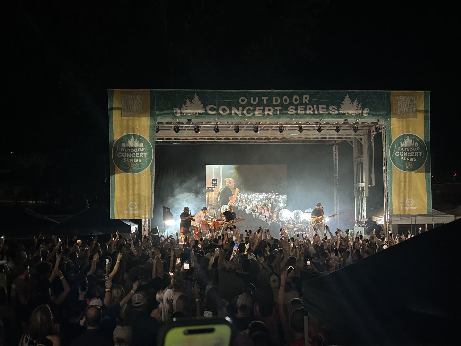 Night crowd with hands up in front of a lit SL100 stage at the Outdoor Concert Series